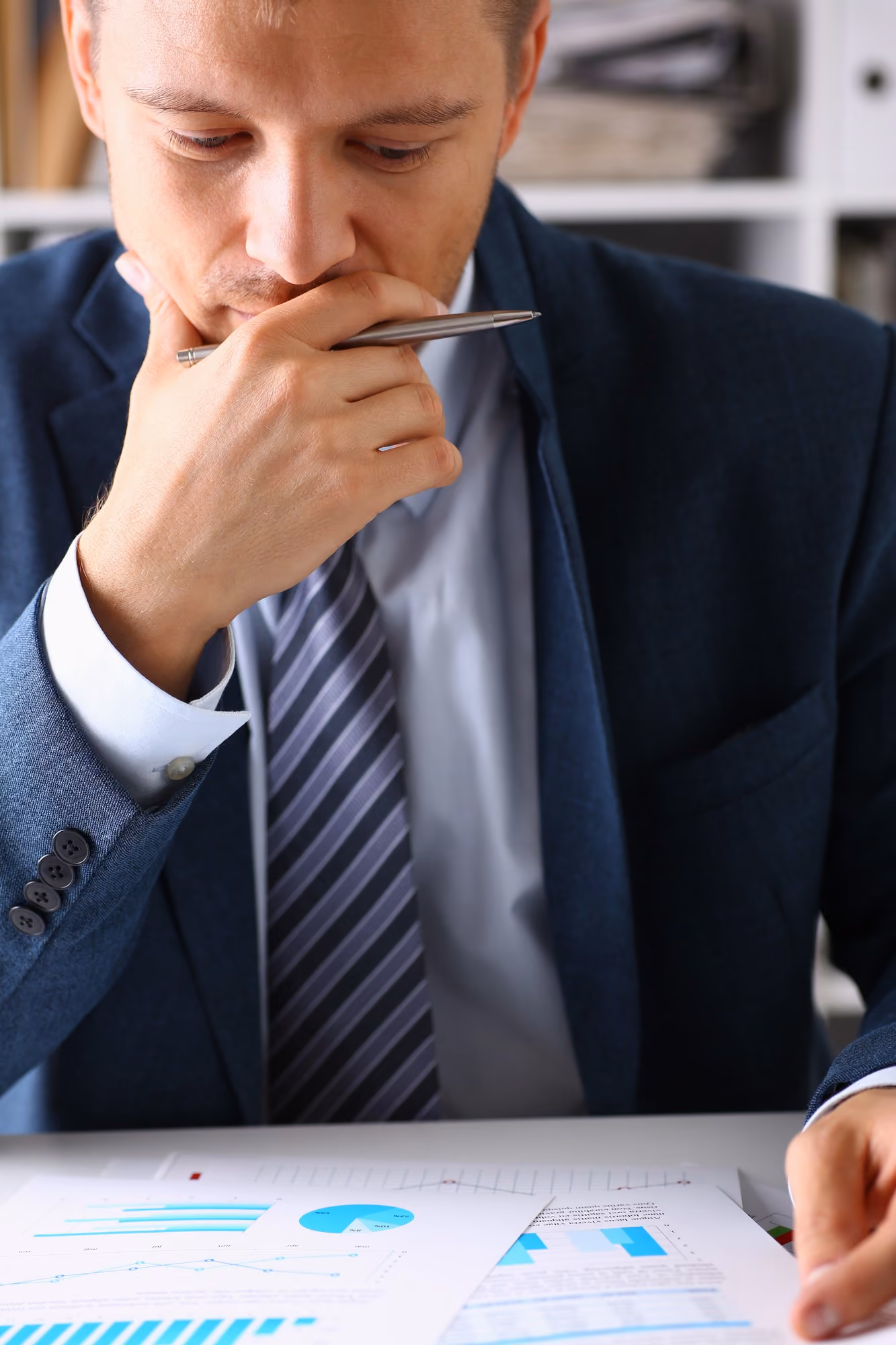 A man in a suit and striped tie holding a pen to his mouth looks thoughtfully at documents and charts on a desk.