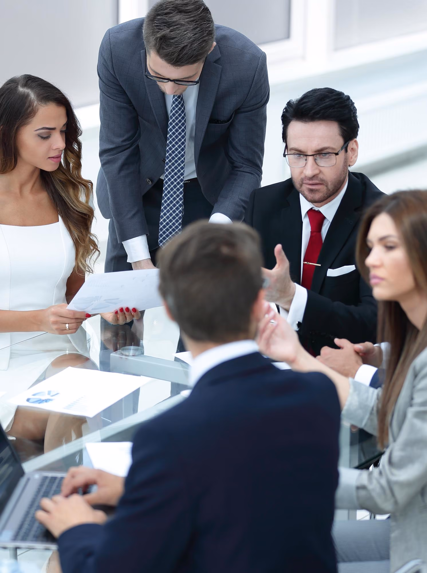 Five business professionals in formal attire gathered around a glass table discussing documents and ideas during a meeting in a bright office.