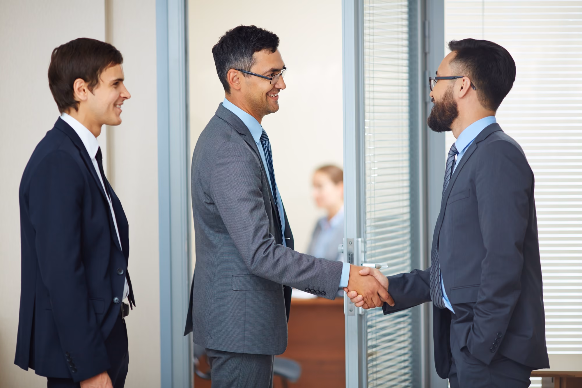 Two men in business suits shaking hands near a glass door while a third man in a suit smiles and watches in an office setting.