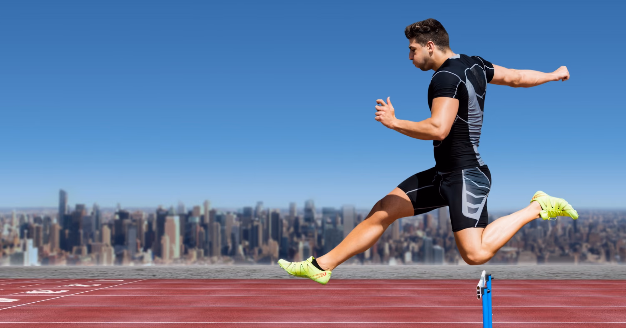 Athletic man in black sportswear with neon green shoes jumping over a hurdle on a red track with a city skyline under a clear blue sky in the background.
