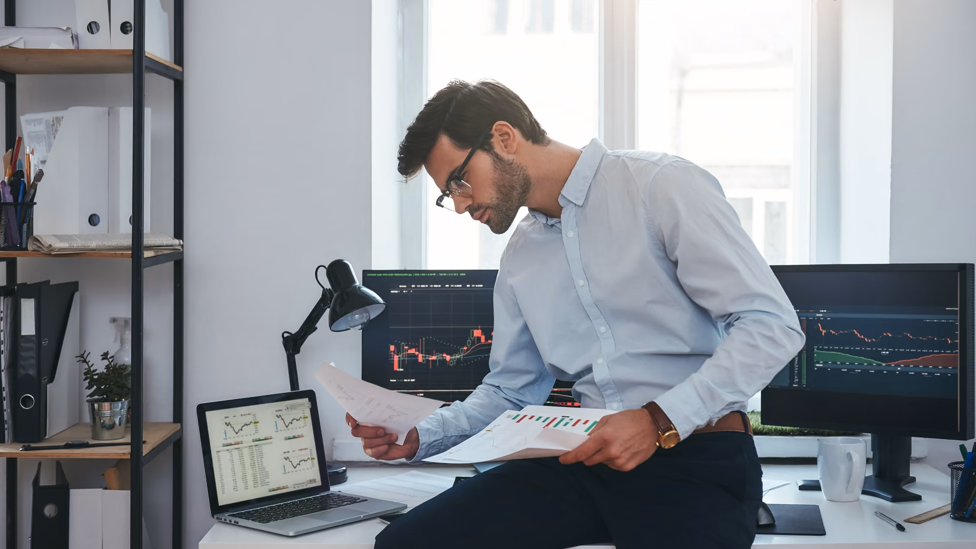 Man wearing glasses and a light blue shirt looking at printed charts while sitting on a desk with a laptop and two monitors displaying stock market graphs in a modern office.