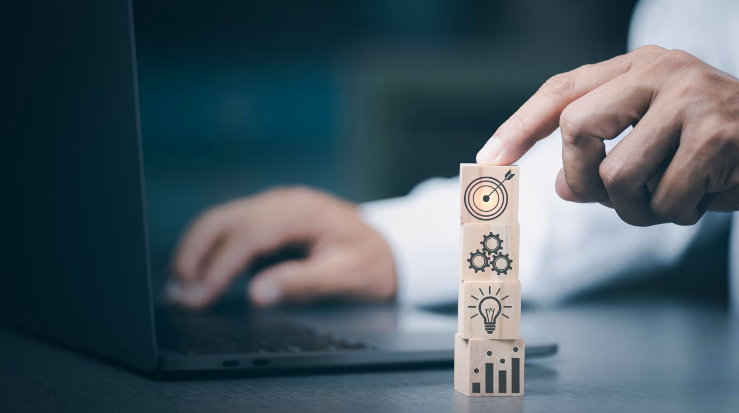 Hand stacking wooden blocks with icons of a bar chart, light bulb, gears, and a target with an arrow, next to a person typing on a laptop.