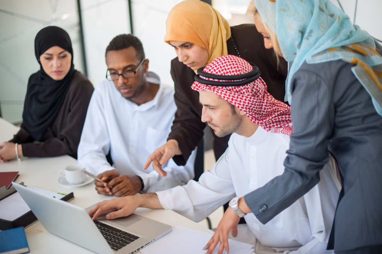 A diverse group of five professionals, including men and women wearing traditional Middle Eastern attire and business clothing, gathered around a laptop engaged in discussion or collaboration.