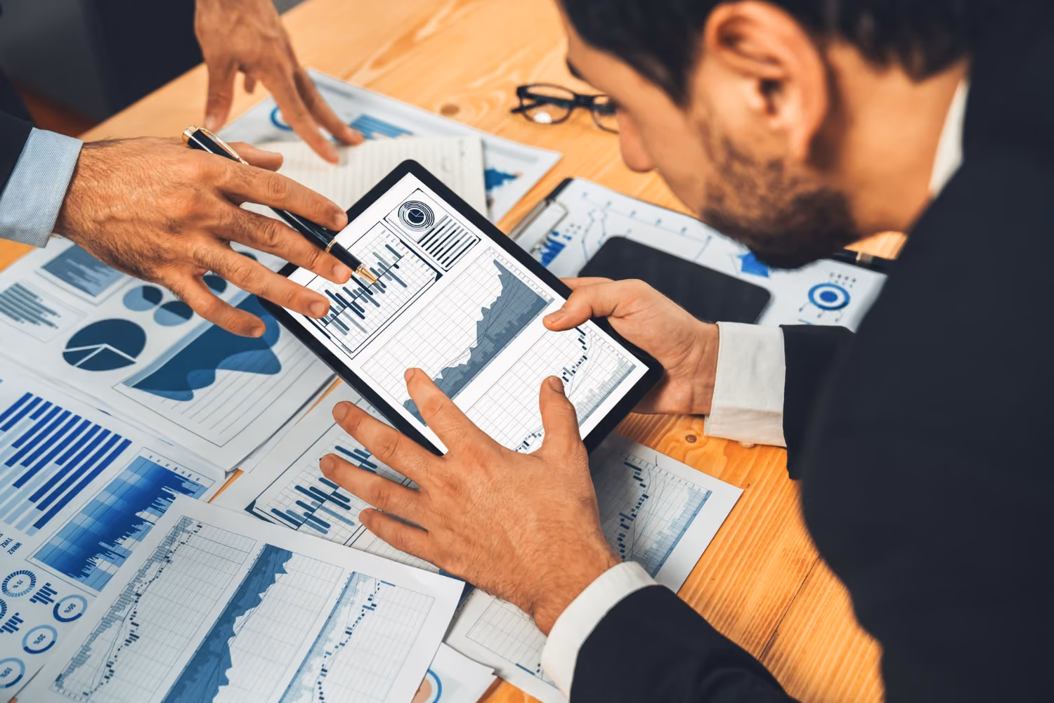 Two business professionals analyzing financial charts and graphs on a tablet and printed documents at a wooden table, discussing data trends.