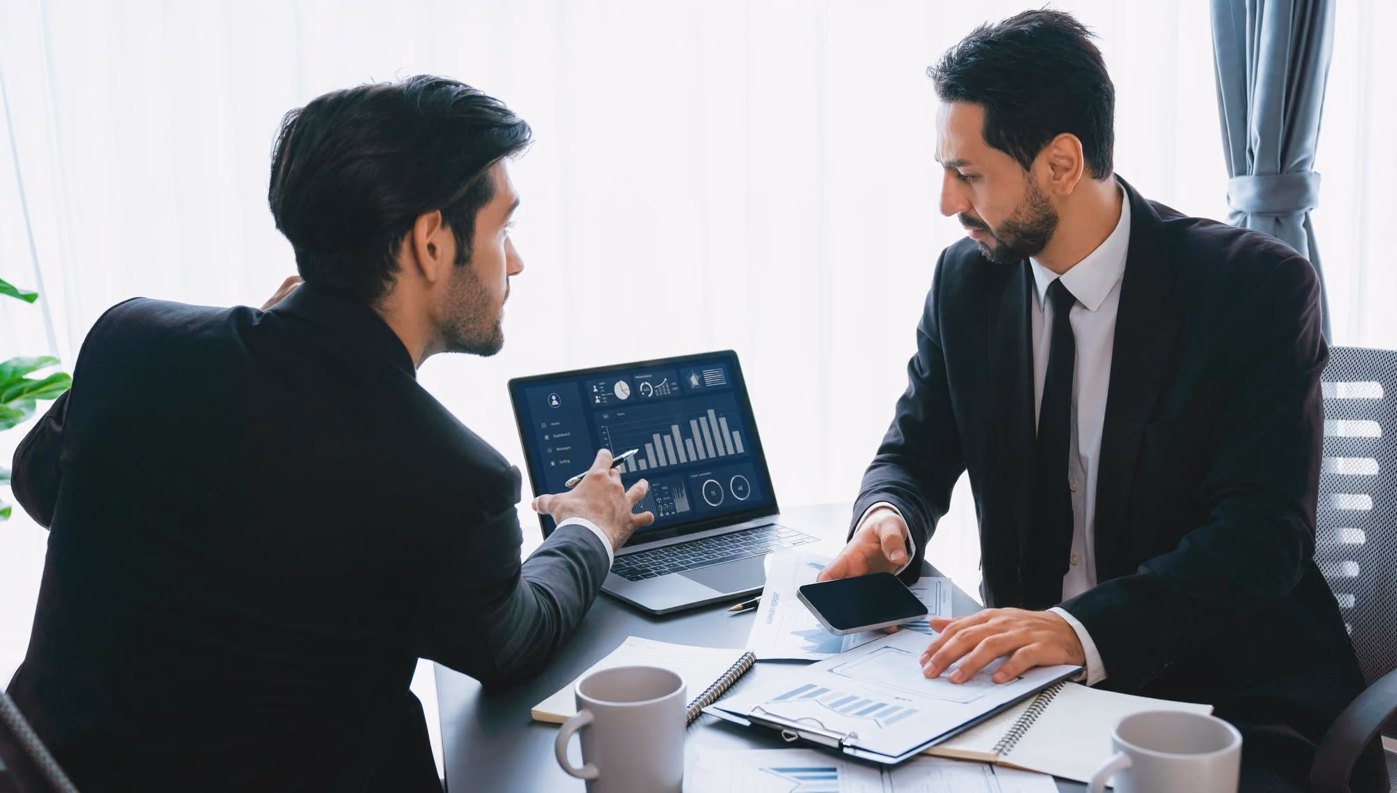 Two businessmen sitting at a table with documents and coffee mugs, discussing data shown on a laptop screen displaying charts and graphs.
