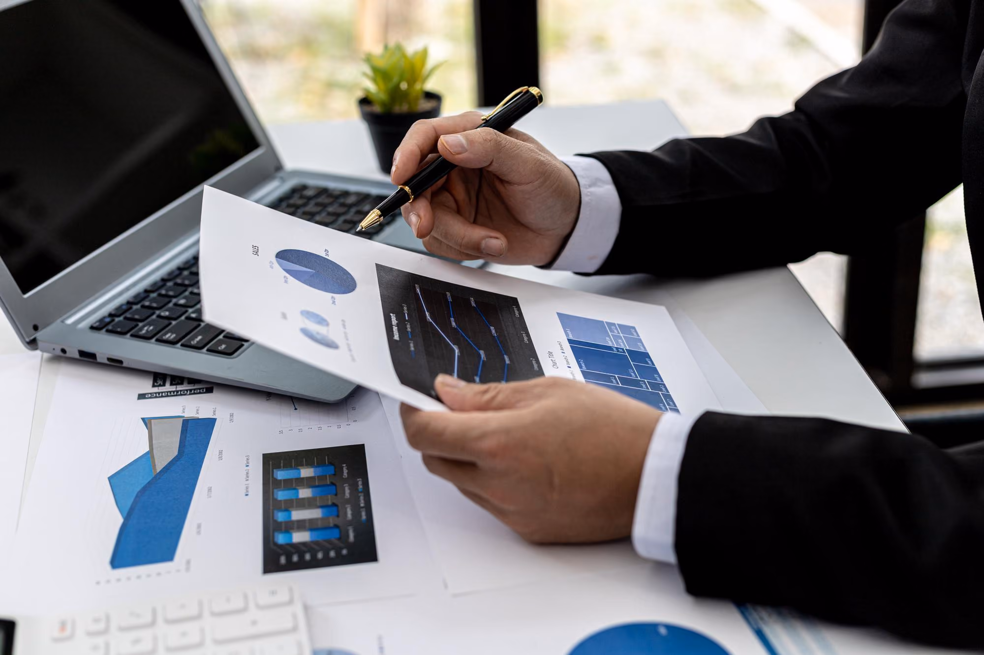 Person in a suit pointing with a pen at charts and graphs on printed papers, with a laptop and a small potted plant on a desk.