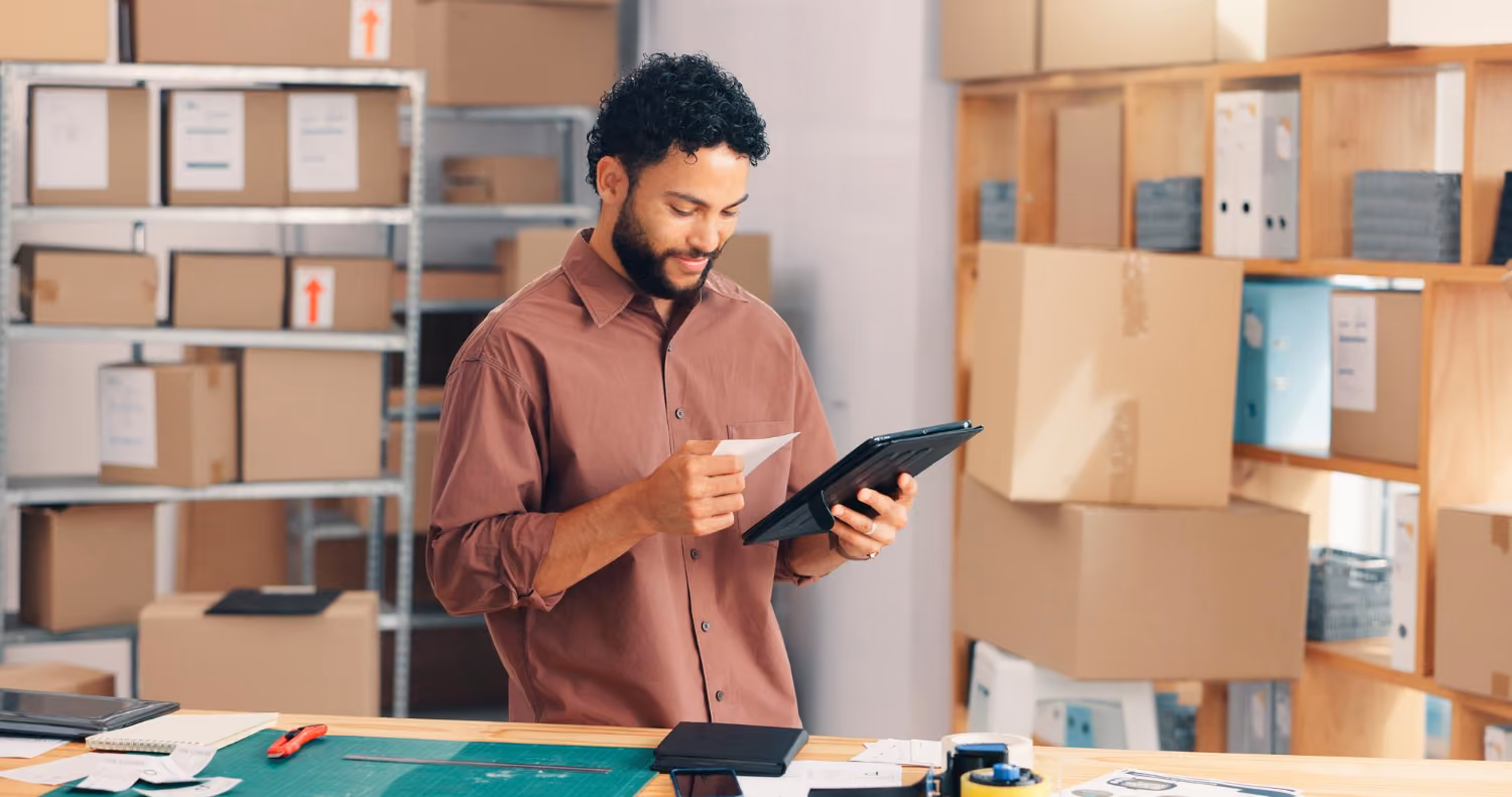 Man with curly hair and beard wearing a brown shirt, checking a receipt and a tablet in a room filled with shelves of cardboard boxes and office supplies.