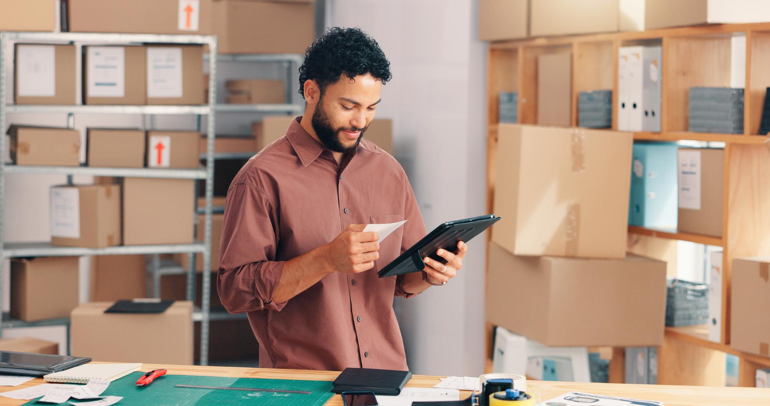 Man with curly hair and beard wearing a brown shirt, checking a receipt and a tablet in a room filled with shelves of cardboard boxes and office supplies.