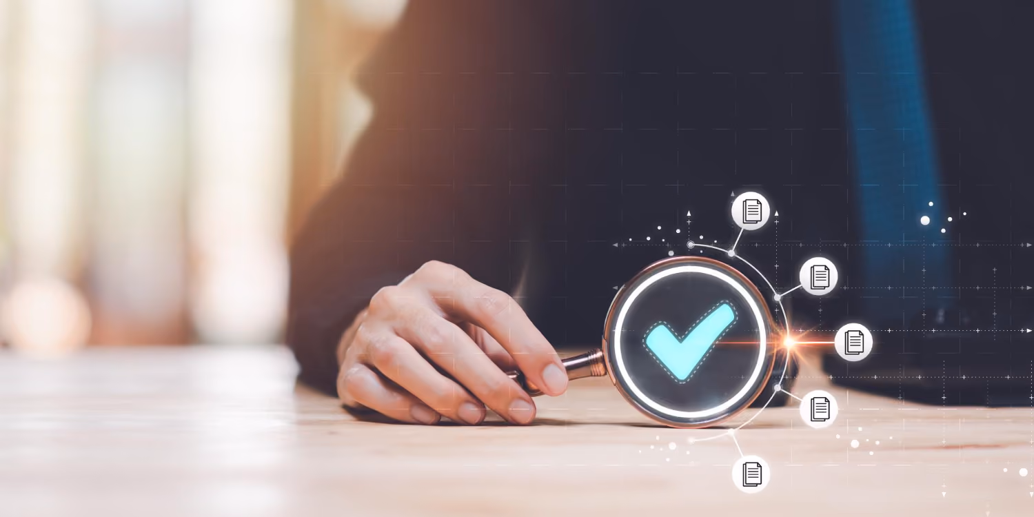 A hand holding a magnifying glass over a wooden surface with a glowing blue checkmark visible inside and various document icons connected around it, symbolizing verification or approval.