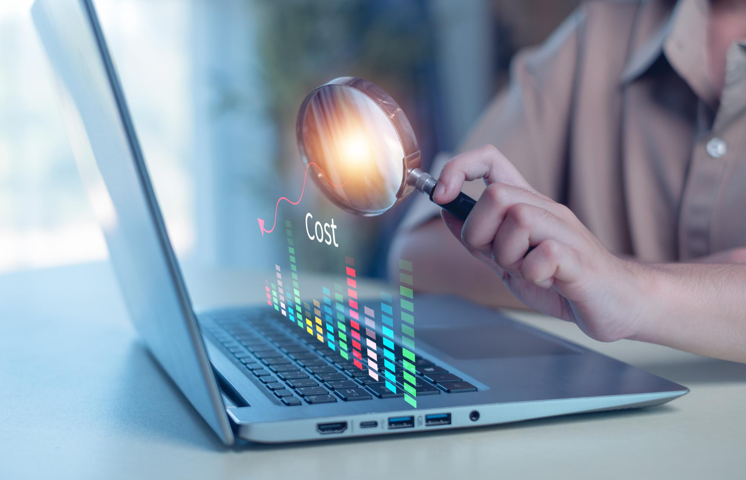 Person holding a magnifying glass over a laptop keyboard highlighting the word 'Cost' and colorful digital bar graph data.