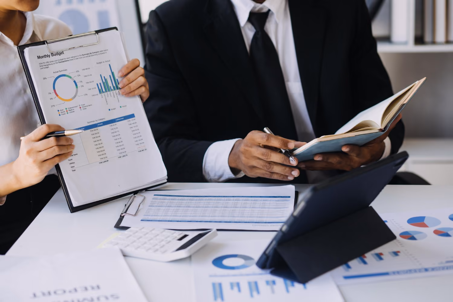 Two professionals at a desk reviewing financial documents; one person holds a clipboard with a monthly budget report featuring pie and bar charts, while the other writes in a notebook. A calculator and tablet are on the desk among additional reports with charts.