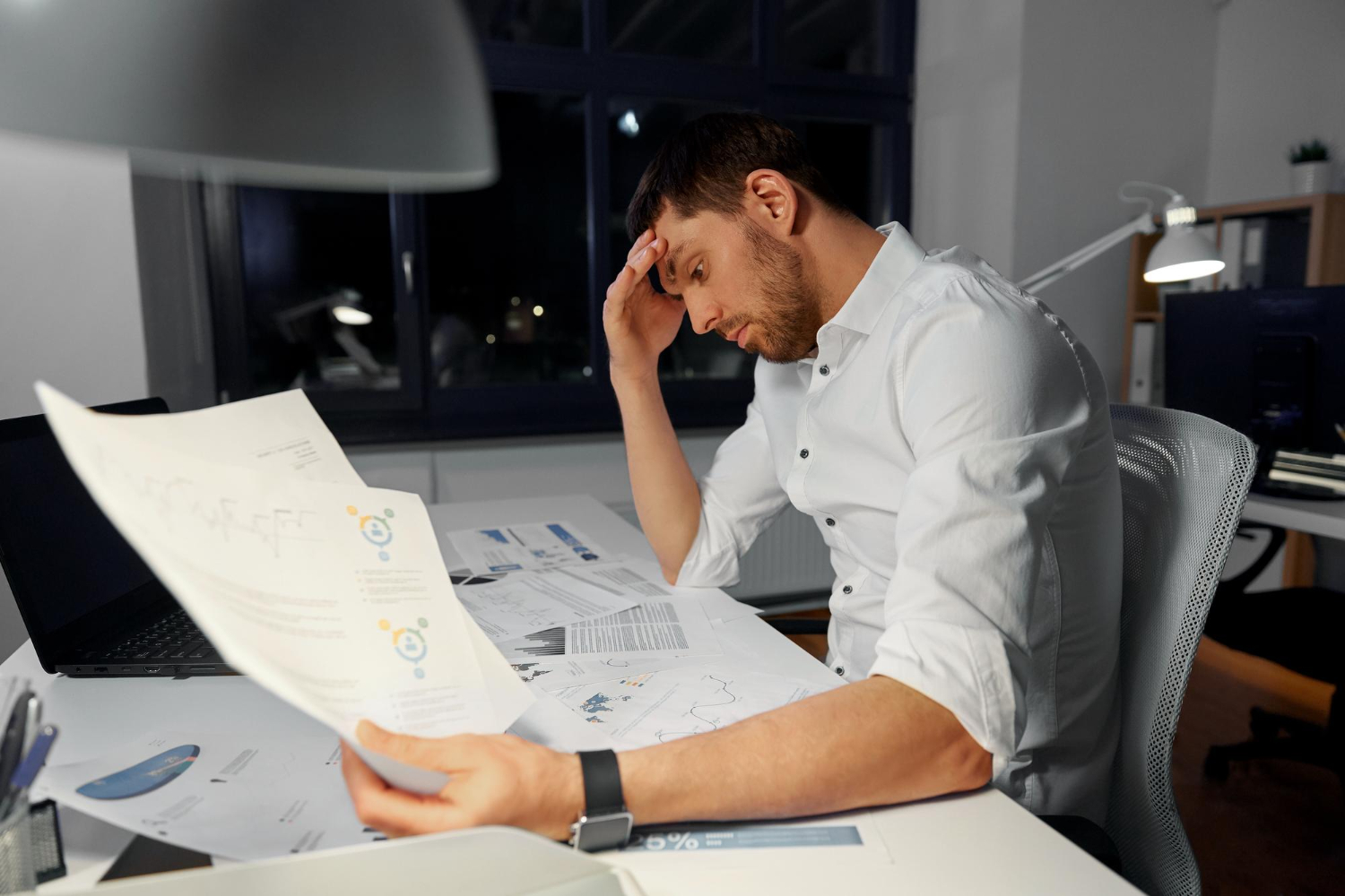 A man in a white shirt sitting at a desk, looking stressed while holding a document and surrounded by multiple papers with charts and graphs in an office at night.