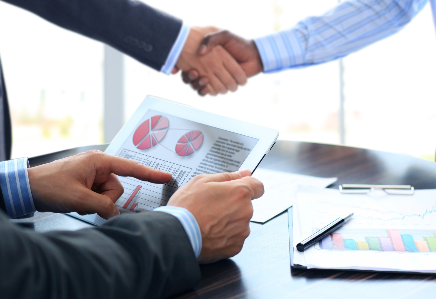 Two people shaking hands over a table while one person holds a tablet displaying red pie charts and graphs, with papers and a pen on the table.