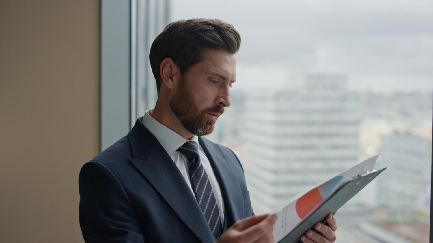 Man in business suit and tie reading a report with colorful pie charts near a window overlooking city buildings.