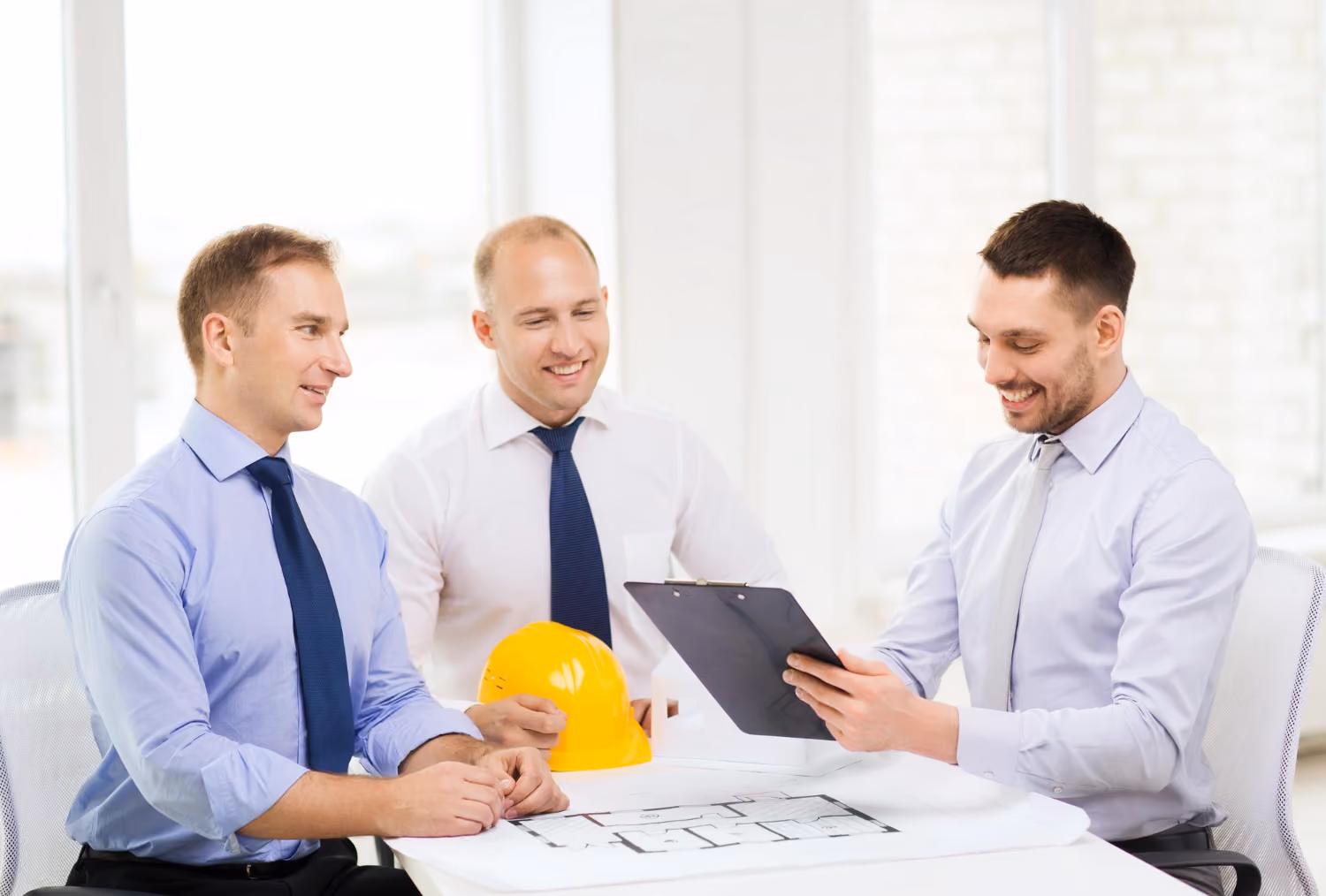 Three men in dress shirts and ties discussing architectural plans at a table, with one man holding a clipboard and another holding a yellow construction helmet.