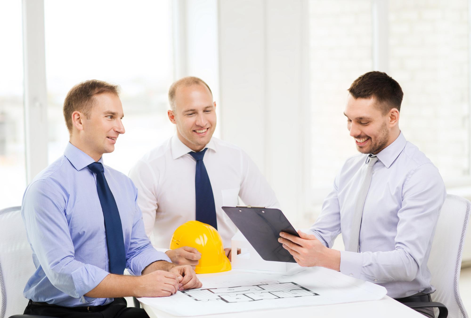 Three men in dress shirts and ties discussing architectural plans at a table, with one man holding a clipboard and another holding a yellow construction helmet.