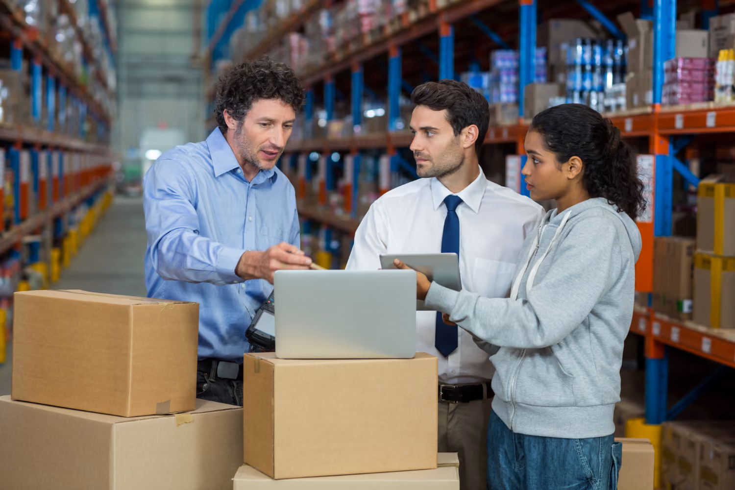 Three warehouse workers standing by stacked cardboard boxes in an aisle of shelves filled with inventory, discussing while looking at a laptop and a tablet.
