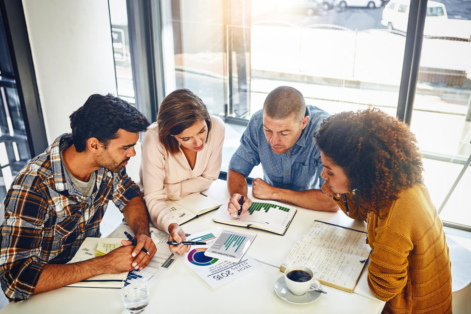 Four diverse colleagues sitting at a table in a bright office, reviewing charts and graphs while discussing data and taking notes.