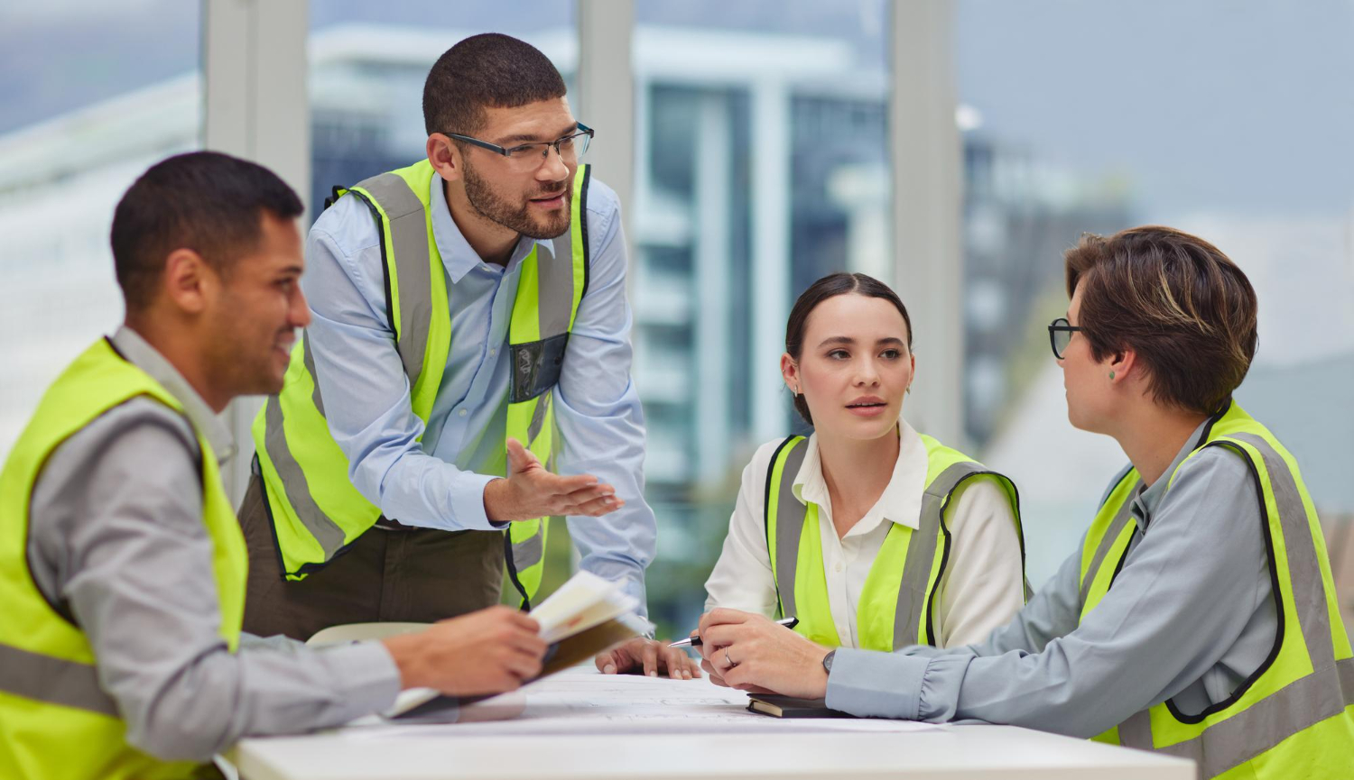 Four construction professionals wearing yellow safety vests having a discussion around a white table with documents and notebooks in a bright office space.