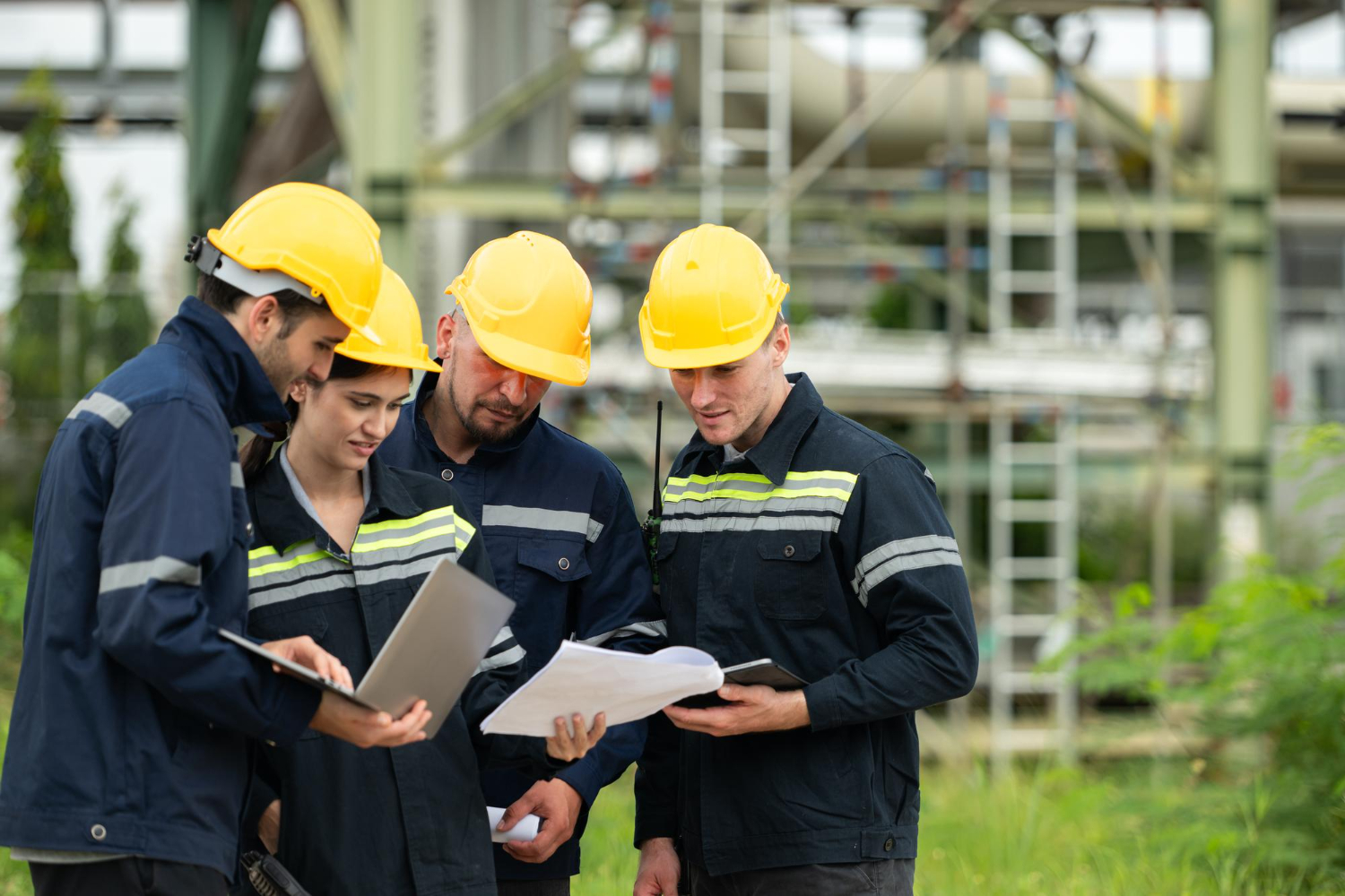 Four construction workers wearing yellow hard hats and navy blue jackets with reflective stripes stand together outdoors, looking at papers and a laptop, with industrial scaffolding in the background.