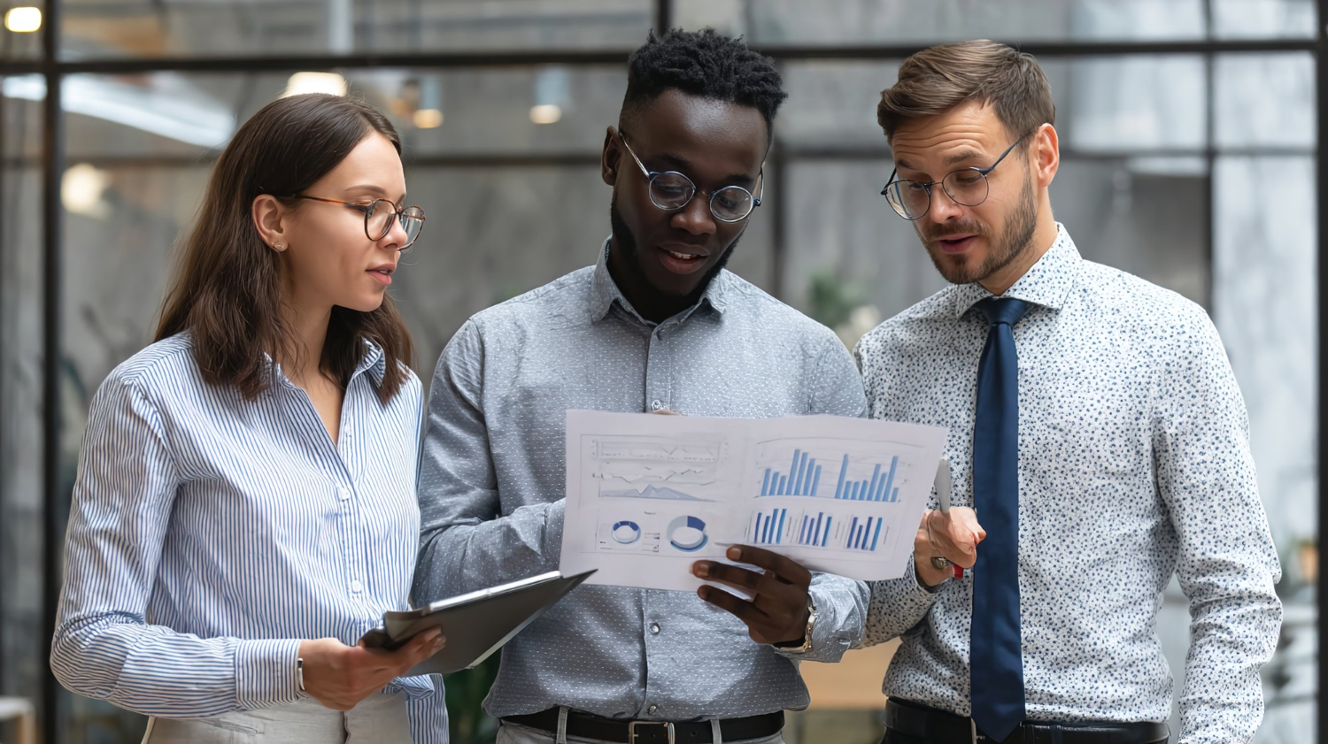 Three diverse professionals standing together in an office, reviewing charts and graphs on printed papers, appearing focused and engaged in discussion.