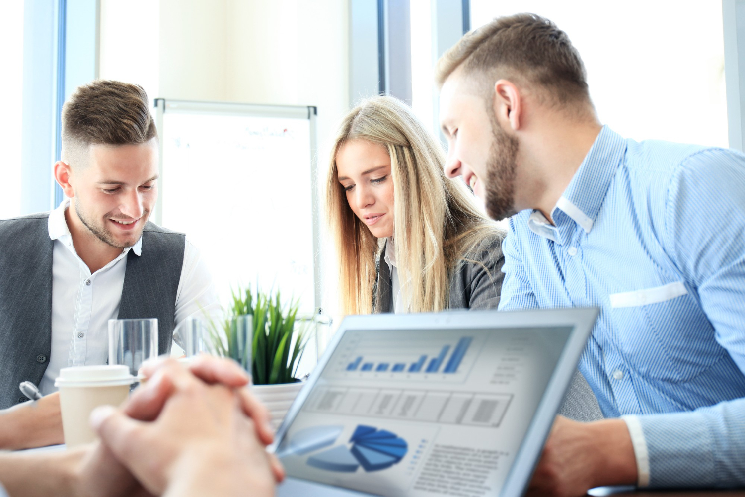 Three young professionals, two men and one woman, seated around a table engaged in discussion with a laptop displaying business charts and graphs in front of them.