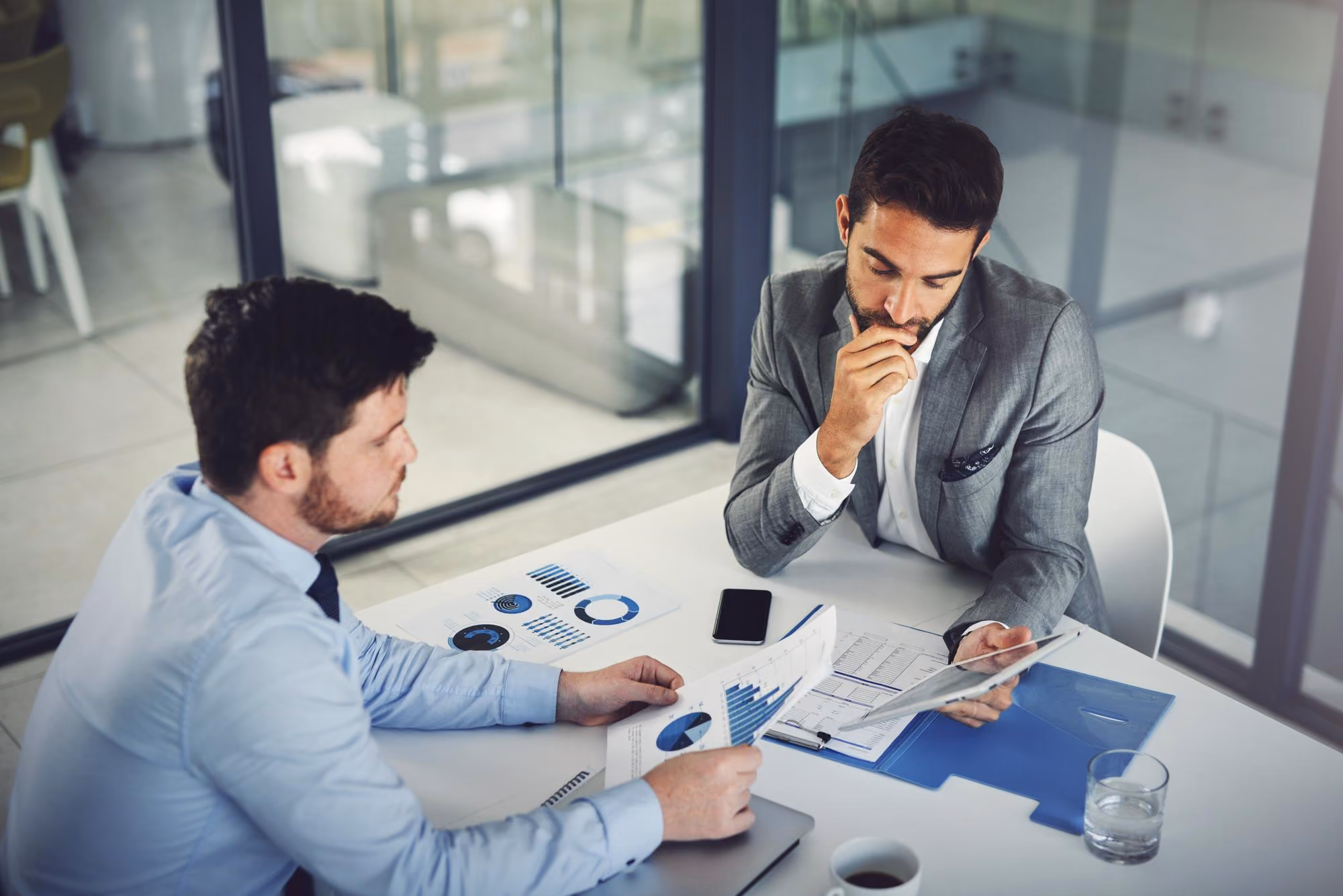 Two men in business attire are sitting at a white table in an office discussing printed charts and graphs, with a smartphone, a glass of water, and a coffee cup nearby.