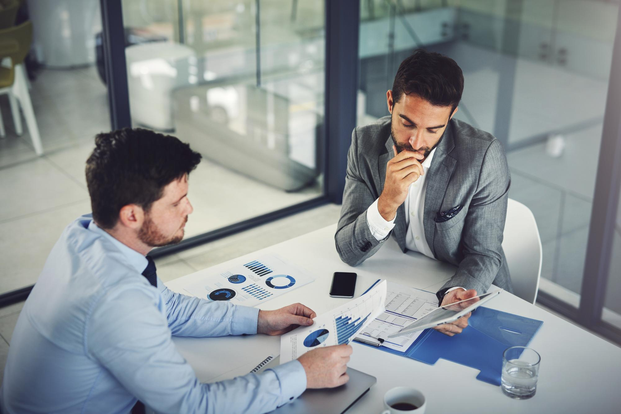 Two men in business attire are sitting at a white table in an office discussing printed charts and graphs, with a smartphone, a glass of water, and a coffee cup nearby.