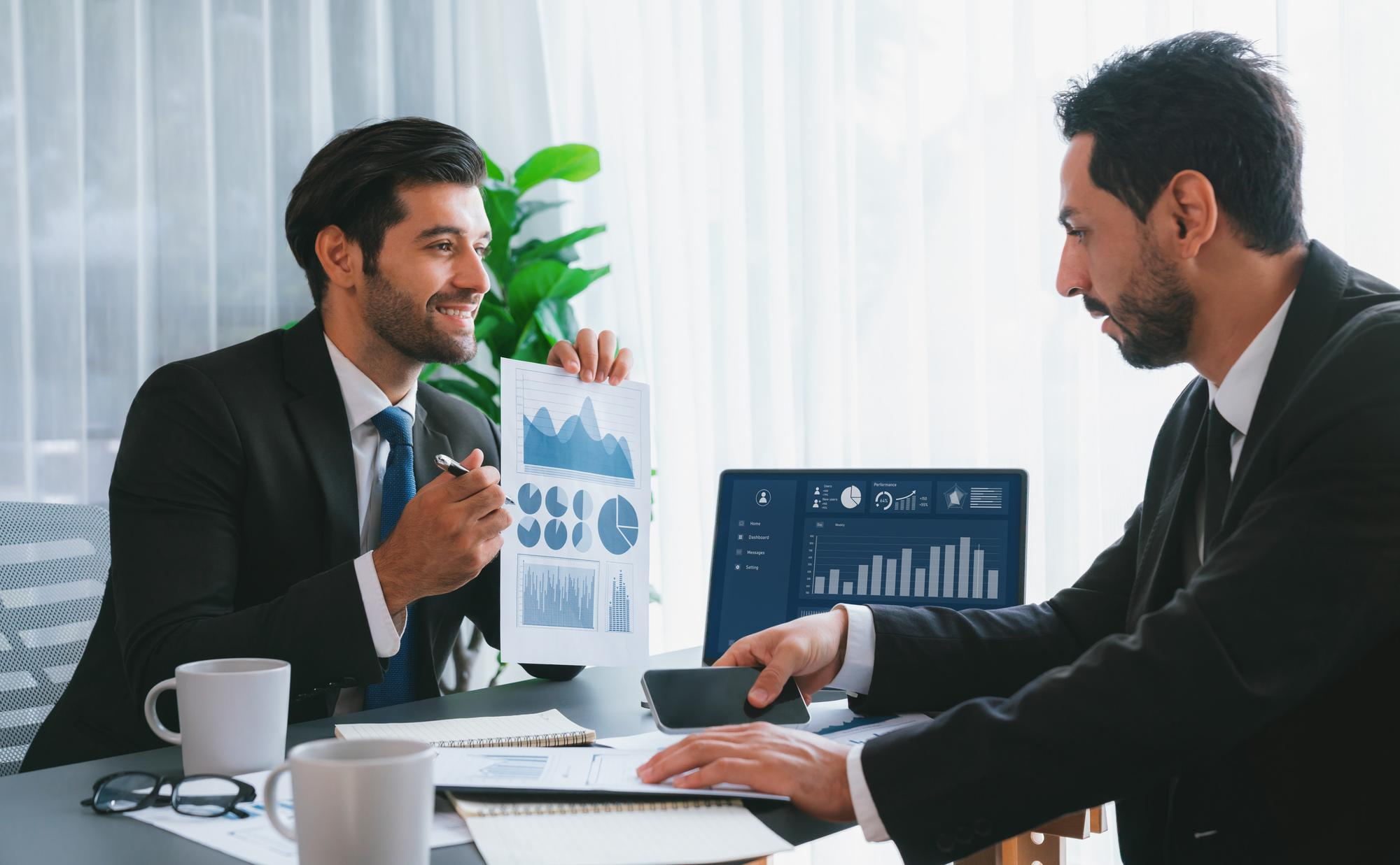 Two businessmen in suits sitting at a table discussing data; one holds a paper with graphs and charts and smiles, while the other looks at the paper, holding a smartphone, with a laptop displaying bar graphs and data in the background.