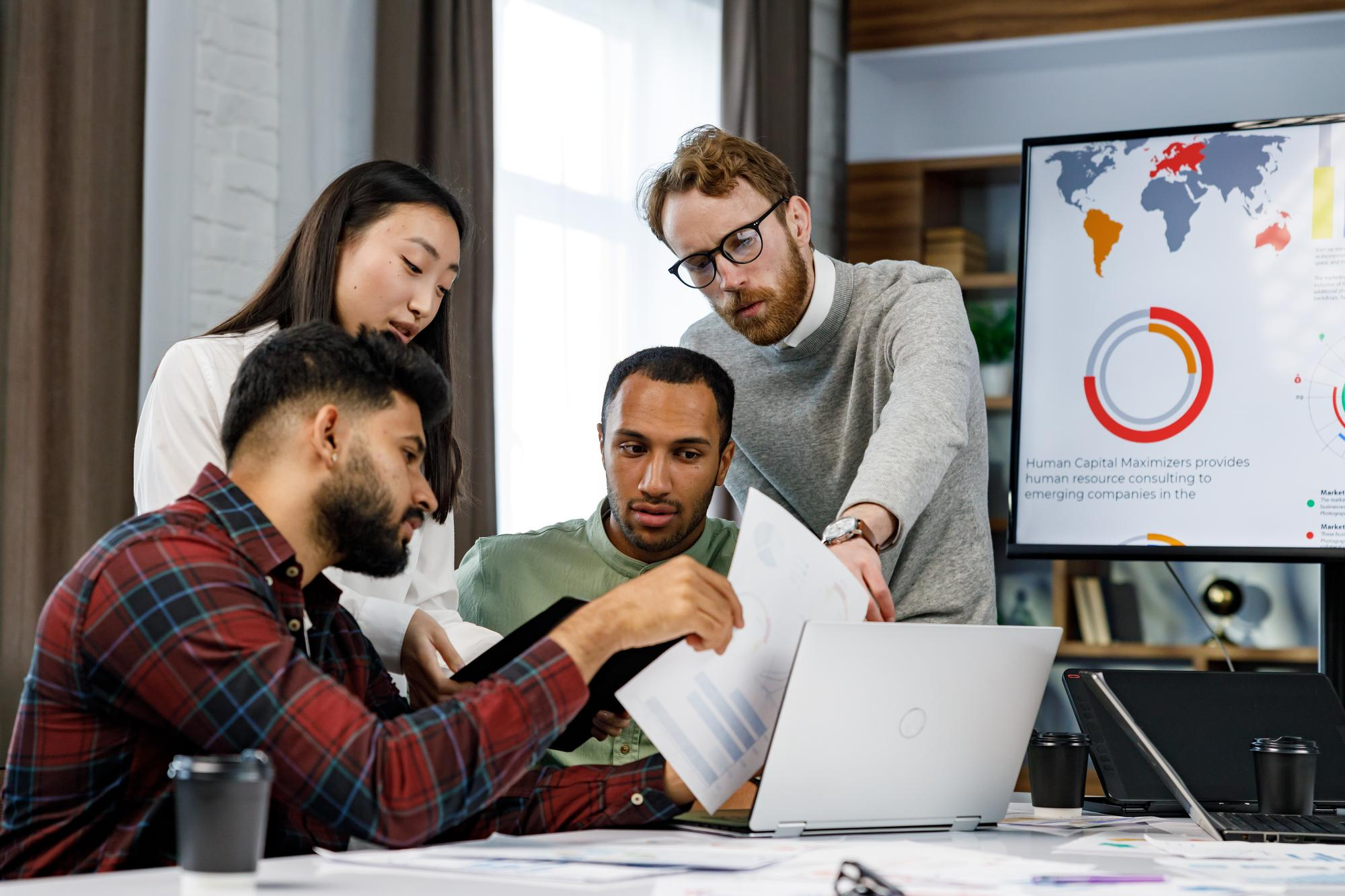 Four diverse colleagues collaborate in a modern office, reviewing charts and data on papers and laptops, with a large screen displaying a world map and graphs in the background.