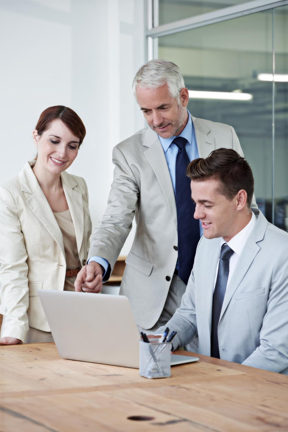 Three businesspeople in light-colored suits gathered around a wooden table looking at a laptop screen, with one man pointing to it while the others smile.