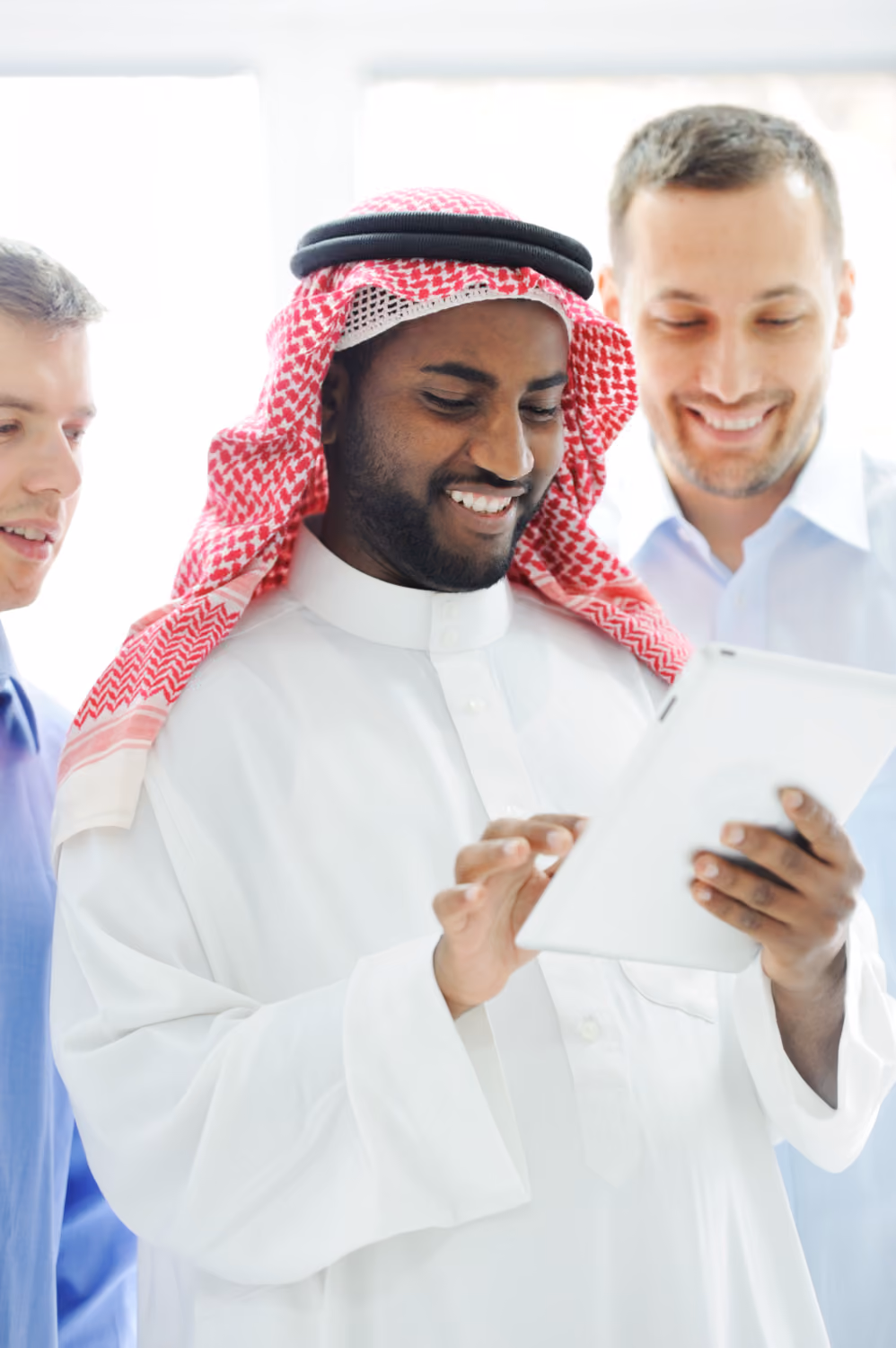 Three men smiling and looking at a tablet, with the man in the center wearing traditional Middle Eastern attire and holding the device.