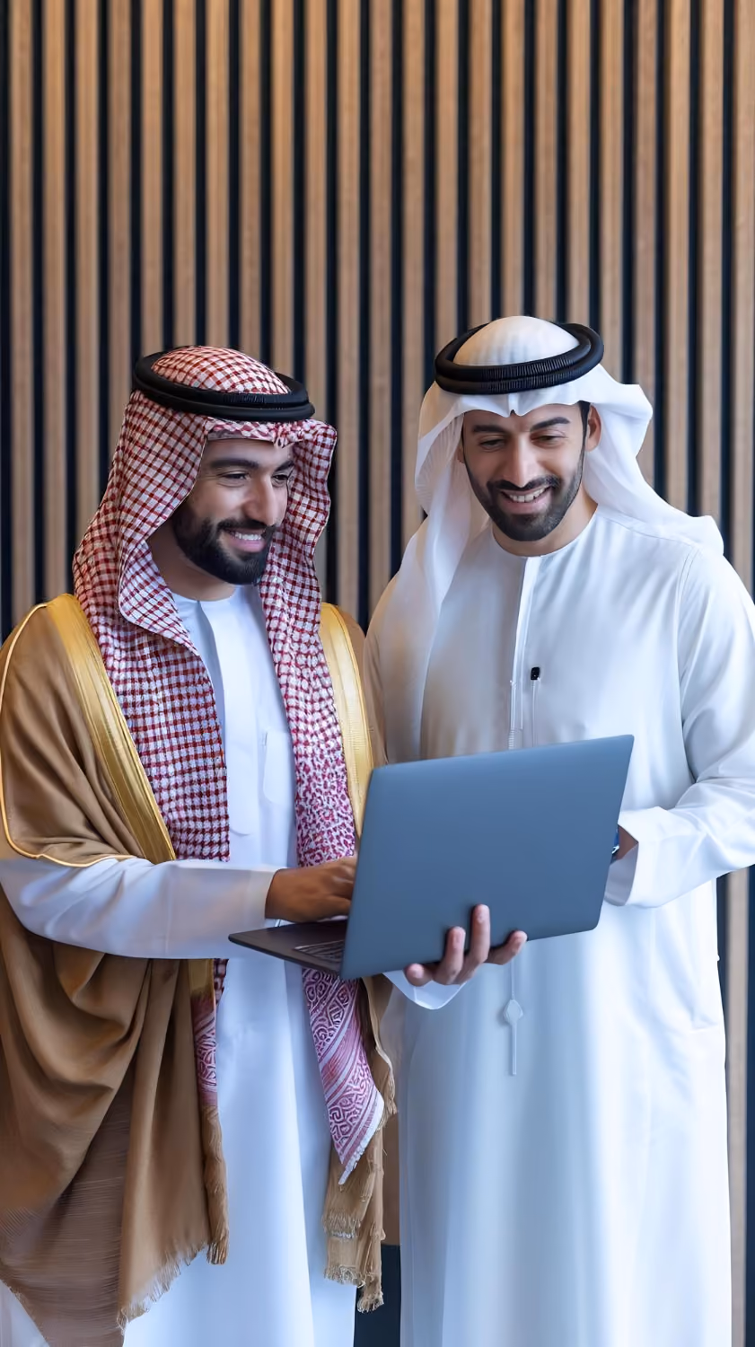 Two Middle Eastern men wearing traditional attire smiling and looking at a laptop together in front of a vertical striped wooden wall.