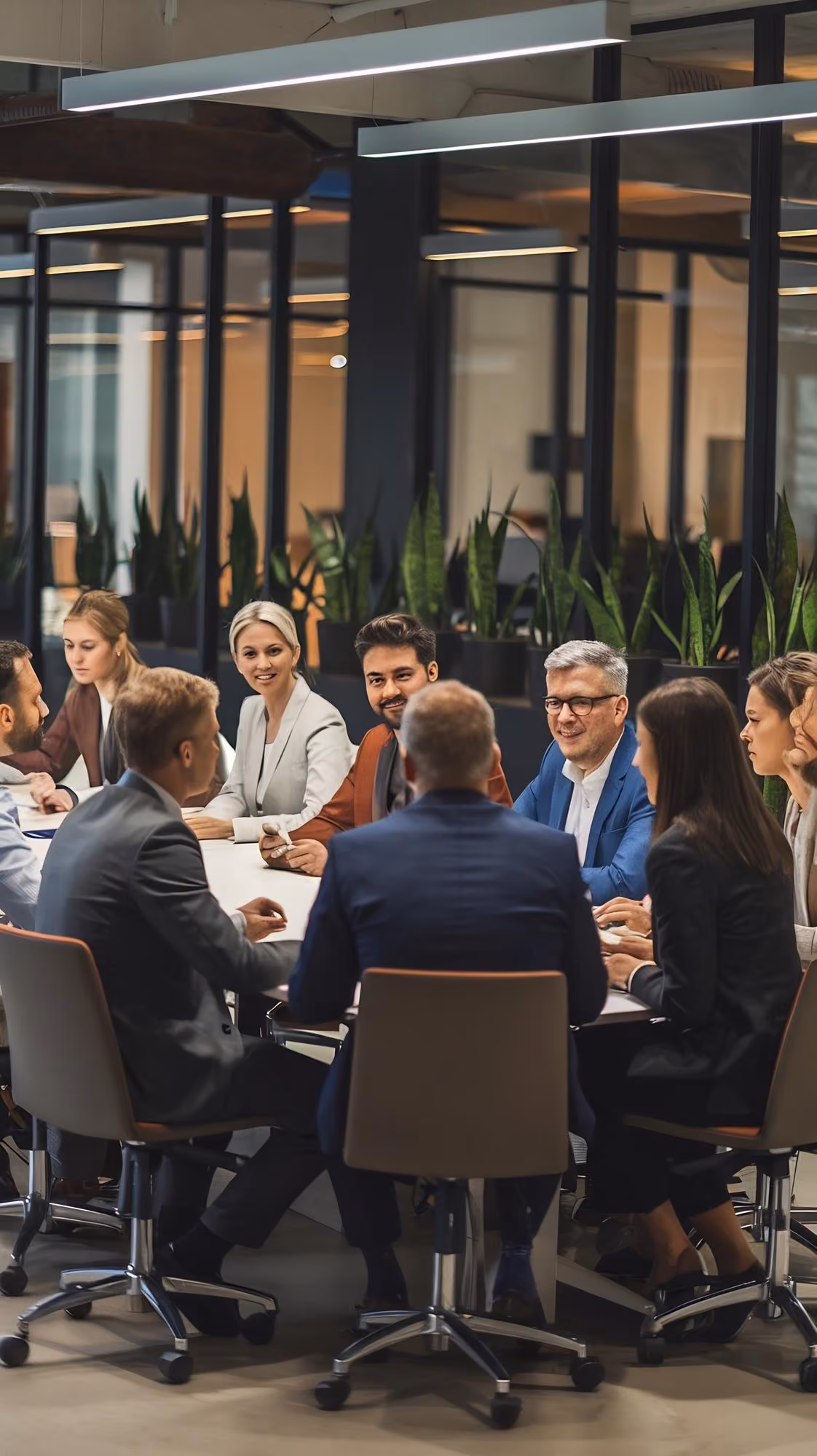 A group of business professionals sitting around a conference table in a modern office having a meeting, with plants and large glass partitions in the background.