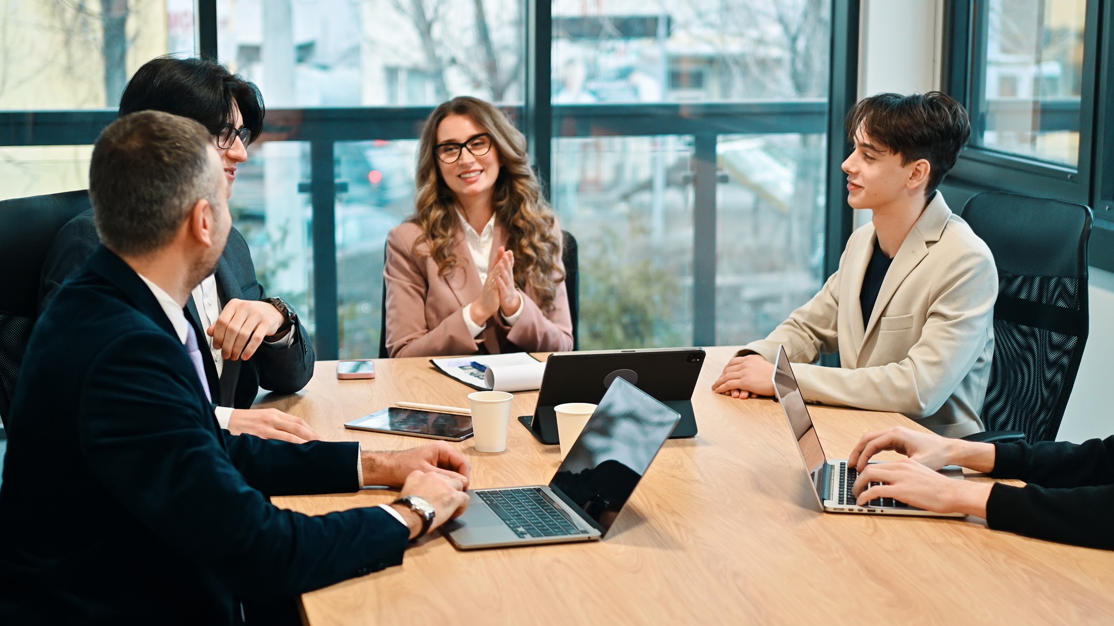Four young professionals having a friendly business meeting around a wooden conference table with laptops, a tablet, notebooks, and paper cups, in a bright modern office with large windows.