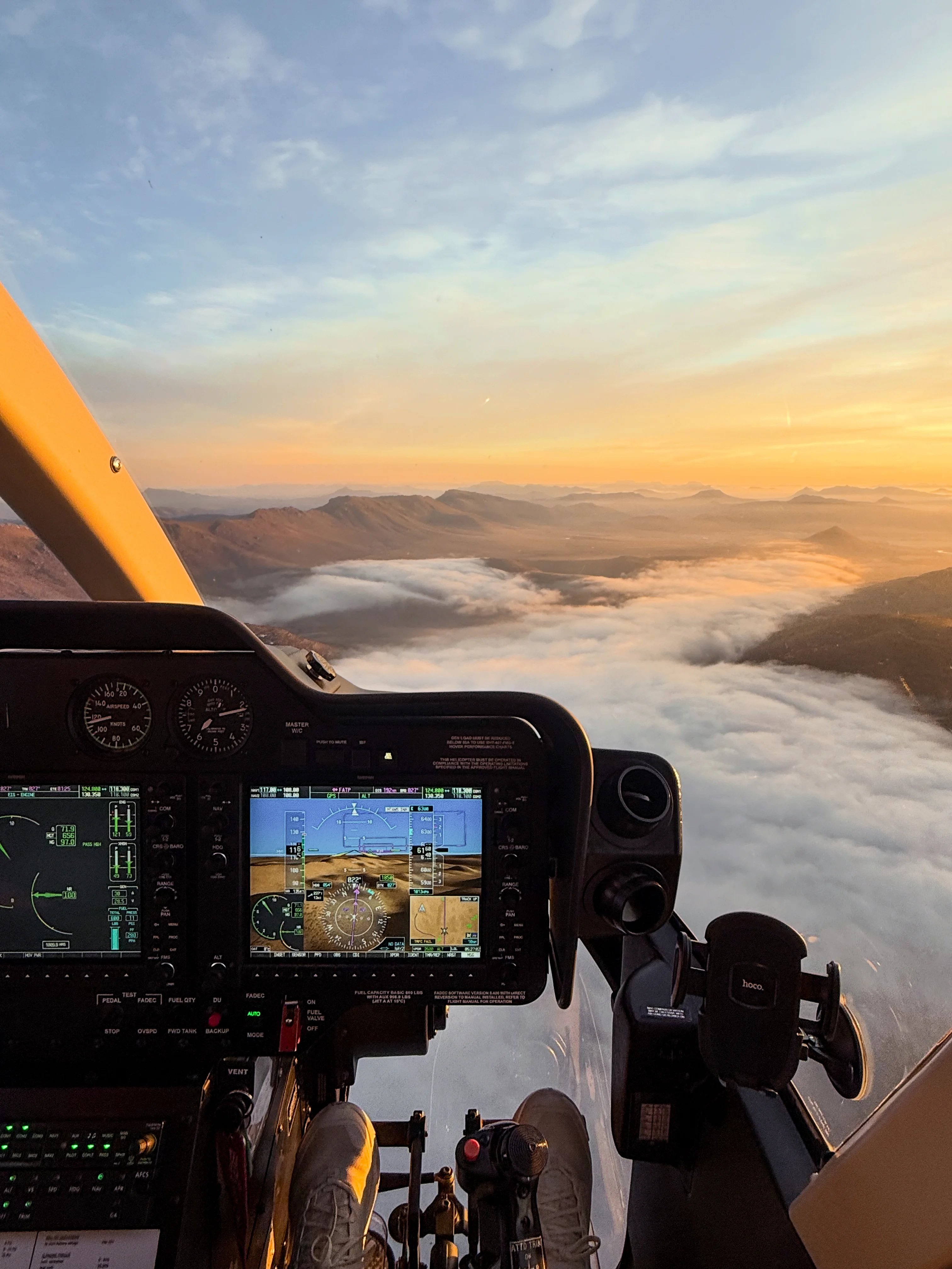 View from helicopter cockpit over clouds and mountain range at sunset with instrument panels visible.