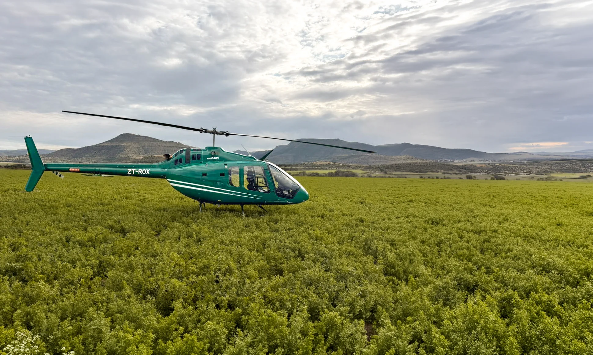 Green Bell 505 helicopter parked in a green field with hills and cloudy sky in the background.