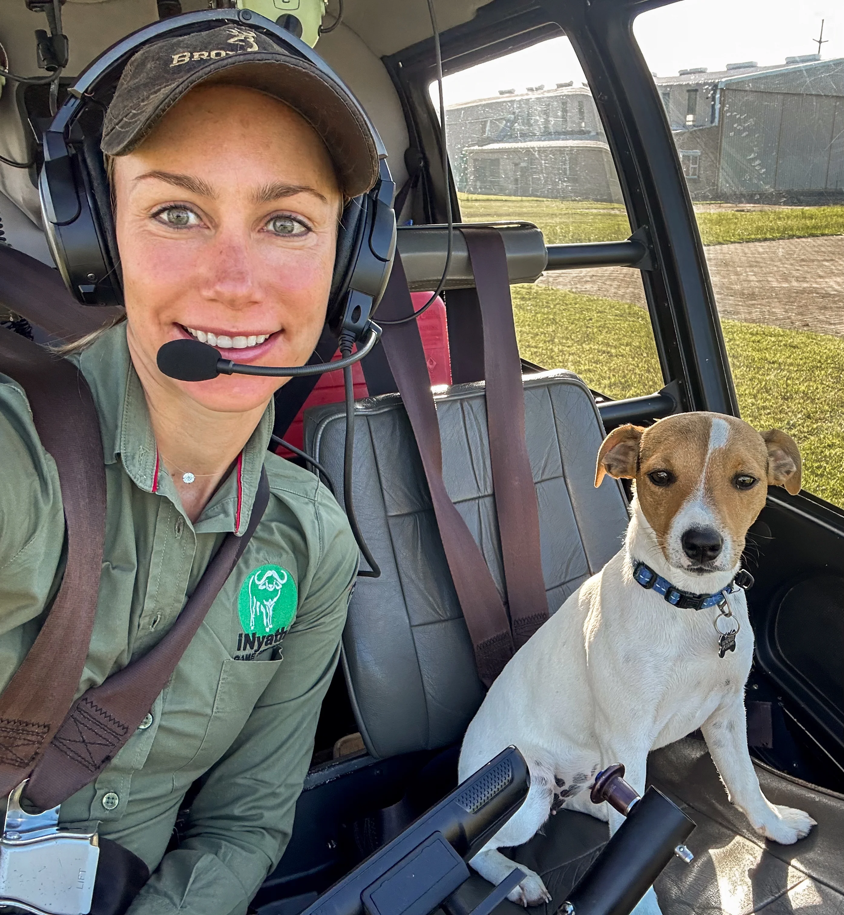 Smiling woman wearing headset and green shirt with dog sitting on a helicopter seat beside her.