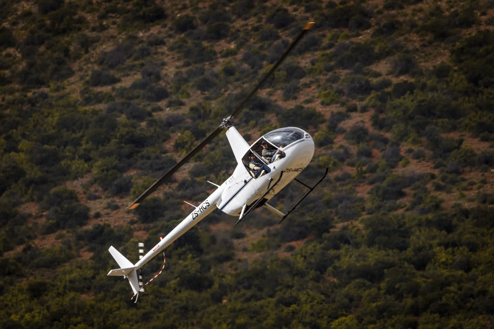 White helicopter in flight with two passengers over a green mountainous landscape.
