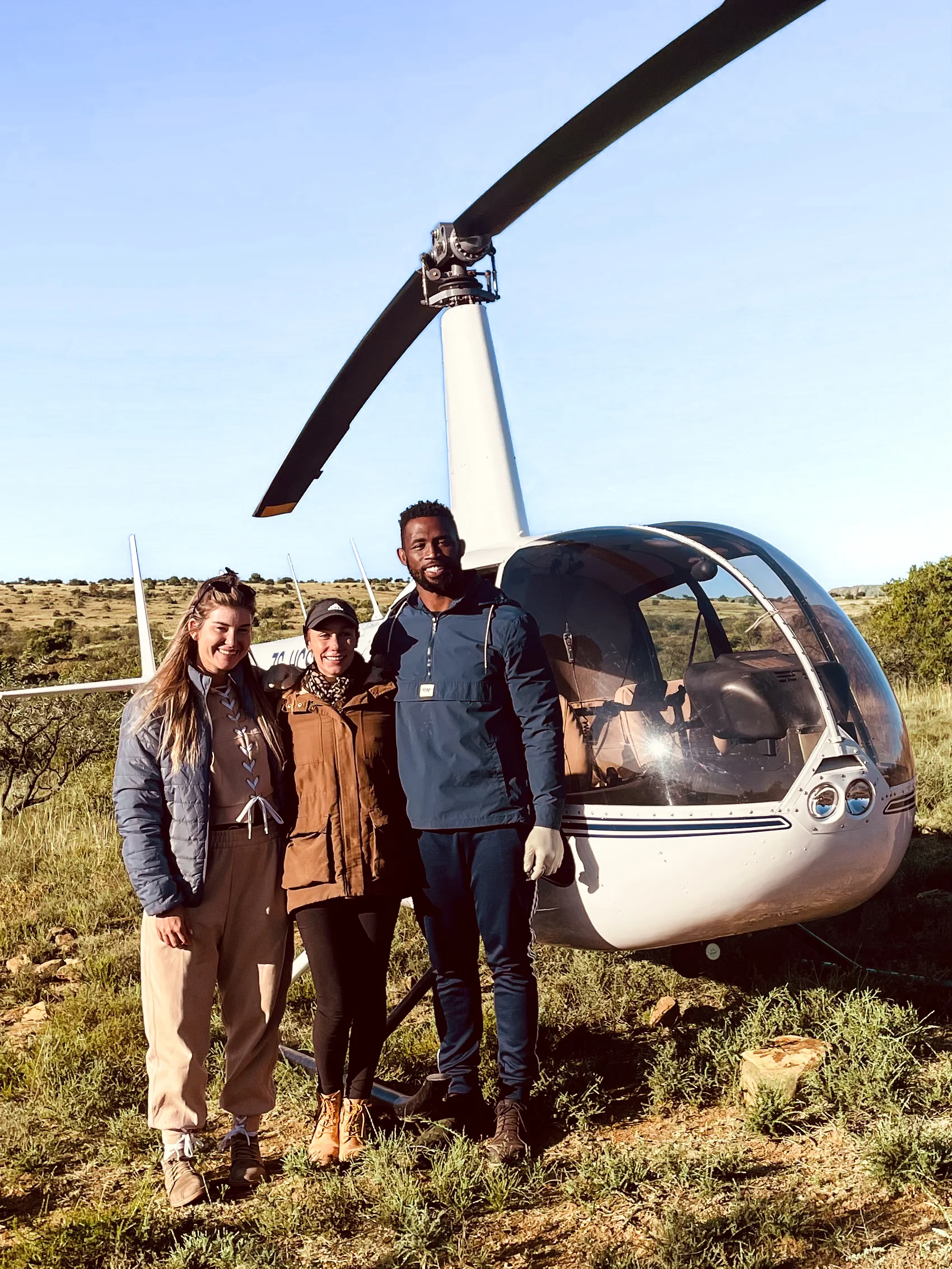 Three adults standing in front of a white helicopter on grass under a clear blue sky.