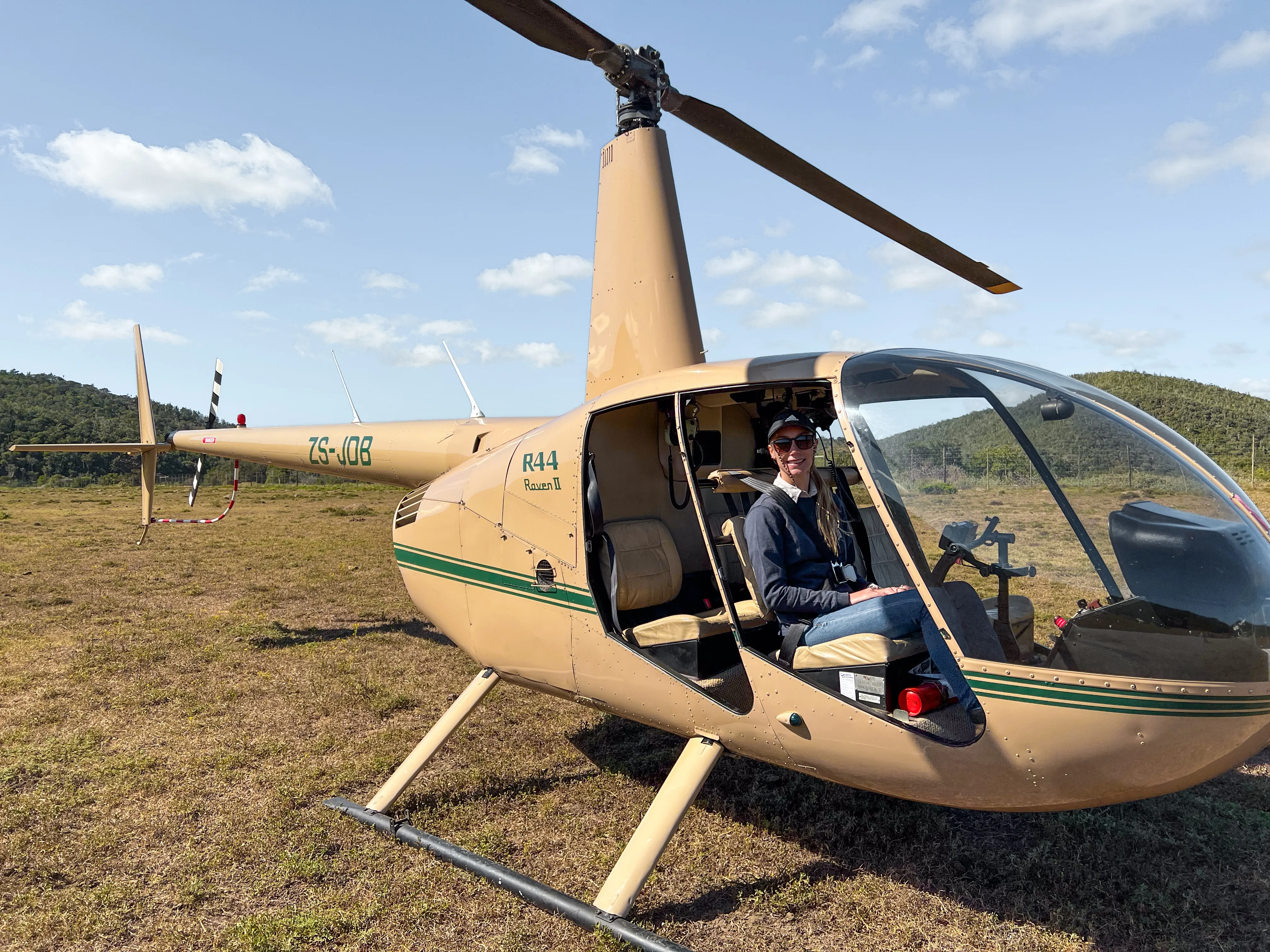 Person seated inside a tan helicopter parked on grassy field with hills and blue sky in the background.