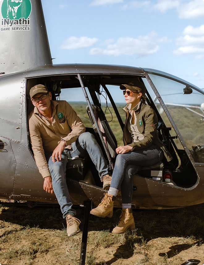 Two people sitting in an open helicopter door wearing iNyathi Game Services clothes in a grassy area.