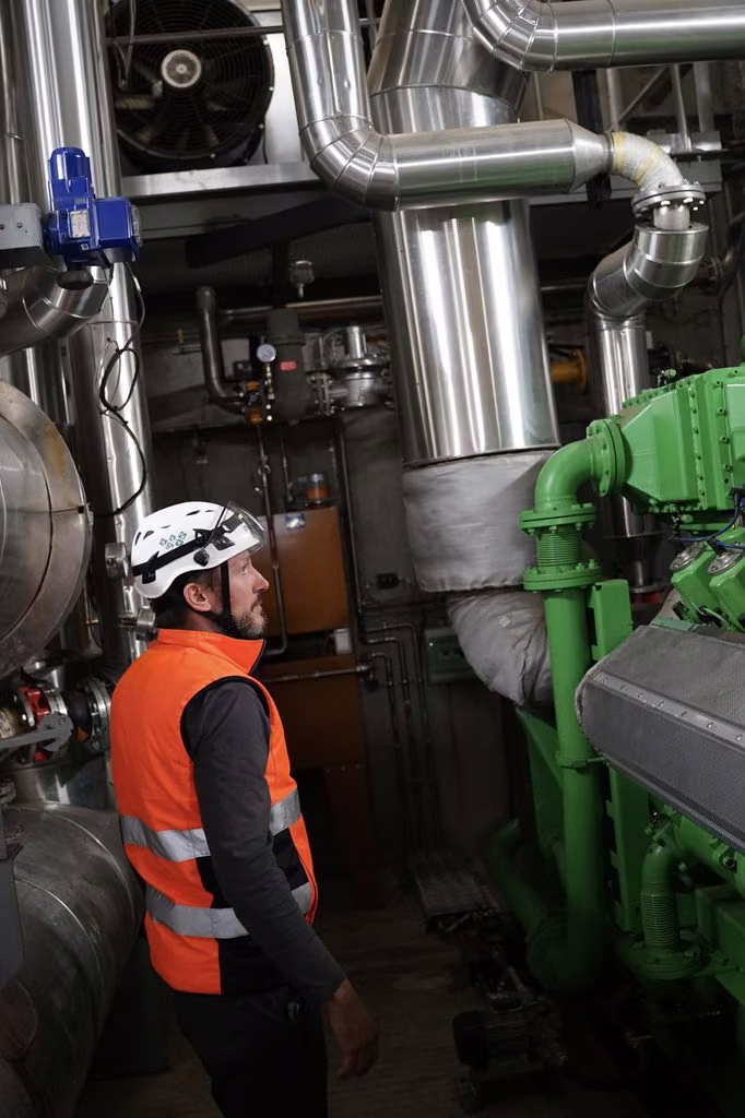 Technicien casqué en gilet orange dans une chaufferie, entre tuyauteries calorifugées et pompes