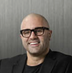 Smiling bald man wearing glasses and a black blazer against a gray background.