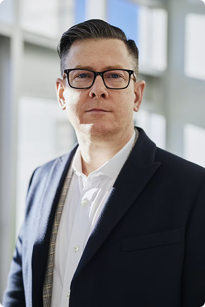 Man with glasses wearing white shirt and dark jacket standing indoors with windows in the background.