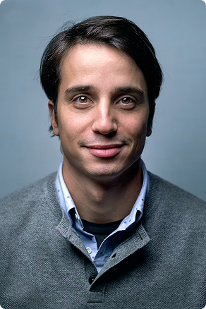 Portrait of a smiling man with short dark hair wearing a grey sweater over a blue collared shirt.
