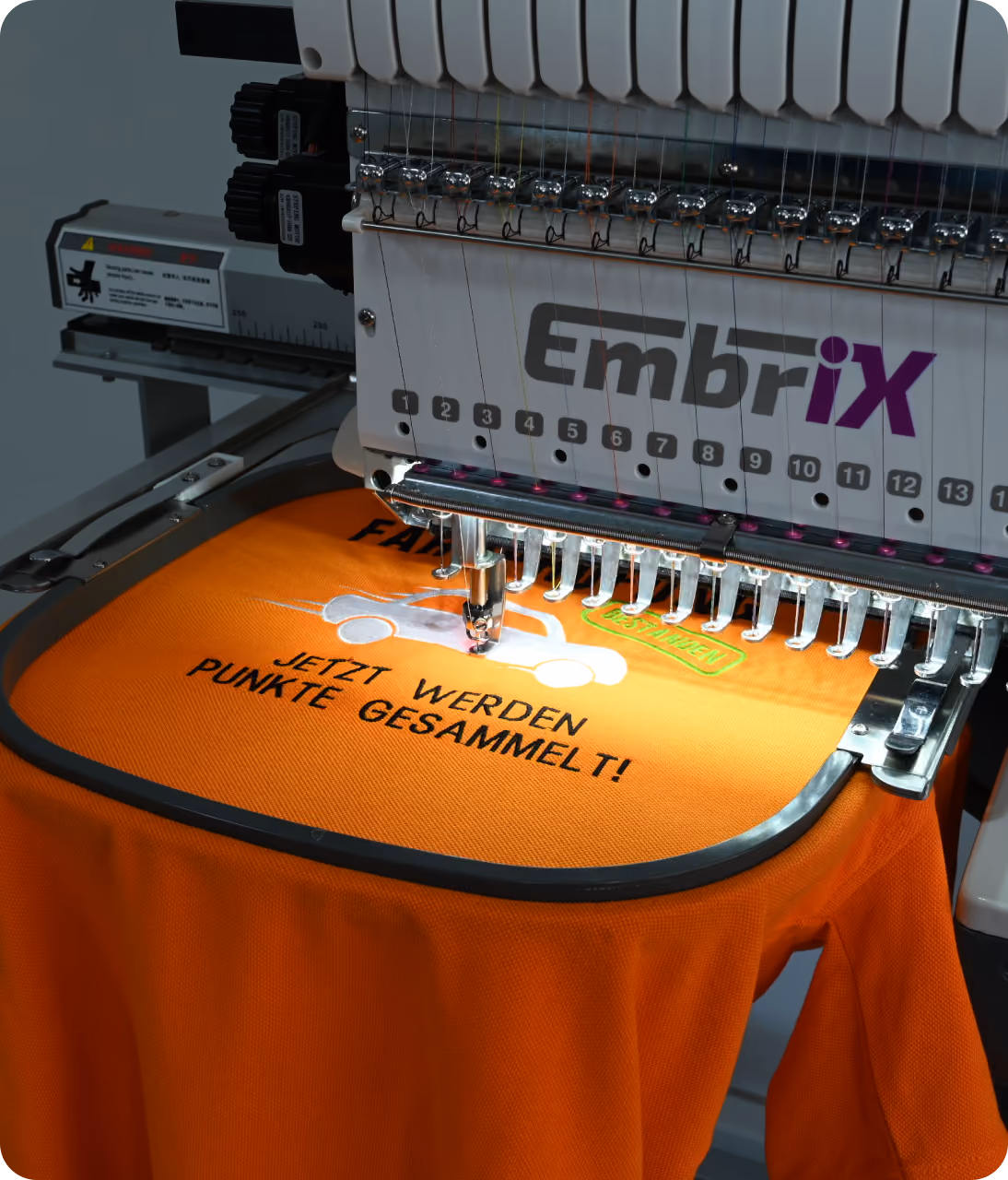 Eine EmbriX-Stickmaschine bestickt ein oranges T-Shirt mit einem Auto und dem Text: Jetzt werden Punkte gesammelt!