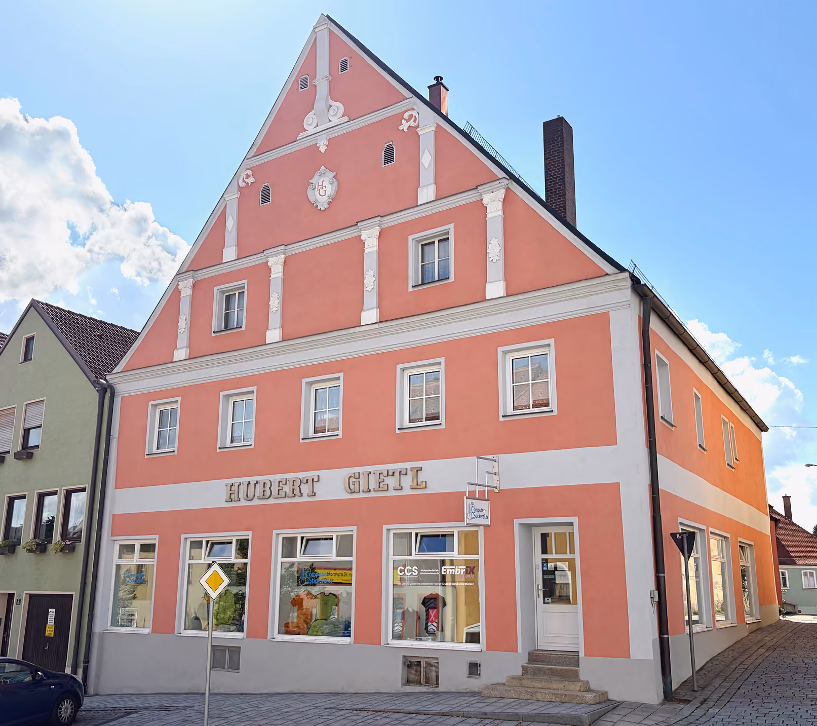 Ein dreistöckiges, rosafarbenes Gebäude mit weißen Verzierungen und großen Fenstern, darauf steht in großen Buchstaben 'Hubert Gietl'. Das Gebäude steht an einer Straßenecke neben einem grünen Haus. Der Himmel ist blau mit einigen Wolken.