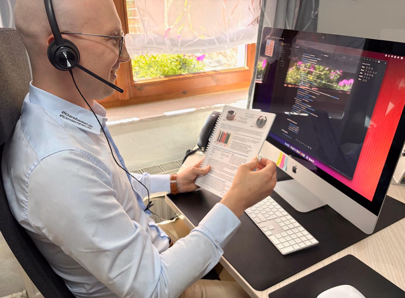 Ein Mann mit Headset sitzt an einem Schreibtisch vor einem iMac und hält eine spiralgebundene Broschüre, während er lächelt. Im Hintergrund ist ein Fenster mit Blumendekoration zu sehen.