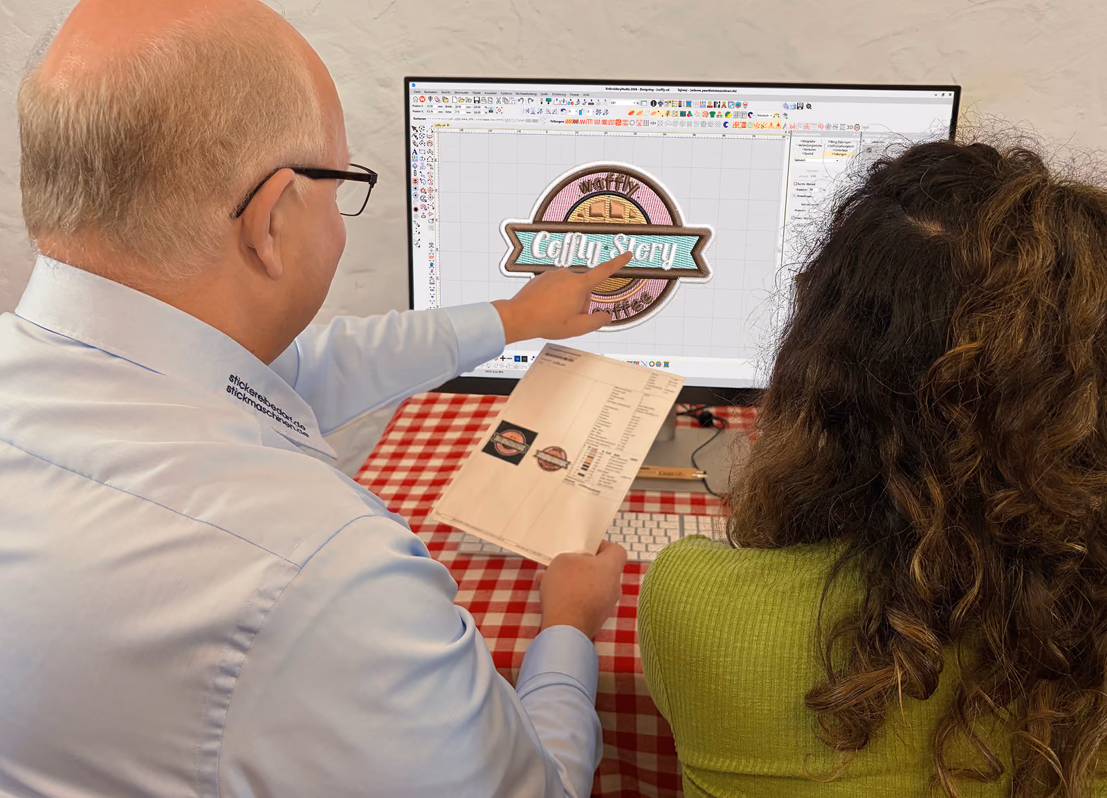 Ein Mann mit Brille zeigt auf ein Computermonitor-Display mit einem digitalen Stickerei-Design, während er einem jungen Mädchen in grünem Pullover gegenüber sitzt und ein gedrucktes Blatt mit Stickerei-Mustern hält.